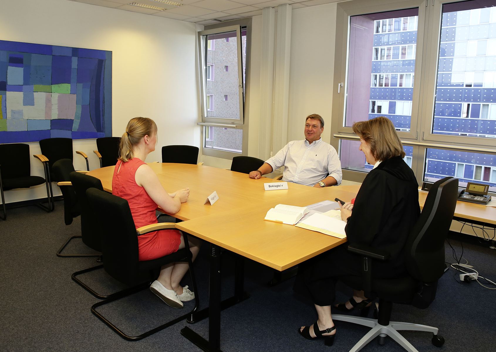 Fotografie des kleinen Sitzungsaals im Verwaltungsgericht Hamburg. In der Mitte des Raumes steht ein großer Besprechungstisch an dem drei Personen sitzen. Am Kopfende sitzt eine dunkel gekleidete Frau mittleren Alters. An der einen Längsseite des Tisches sitzt ein Mann mittleren Alters. Er trägt ein weißes Hemd. Ihm gegenüber sitzt eine jüngere Frau in einem roten Kleid.