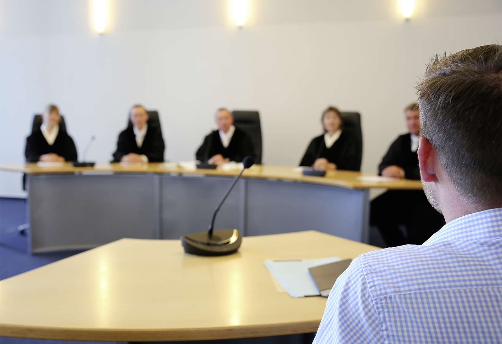 Fotografie des großen Sitzungssaals. Im Hintergrund sitzen in einer Reihe Richterinnen und Richter. Dieser Bereich des Fotos ist unscharf. Vorne rechts im Anschnitt sieht man die Rückenansicht eines jungen Mannes. Er schaut in Richtung der Richterinnen und Richter. Dieser Bereich des Fotos ist scharf.